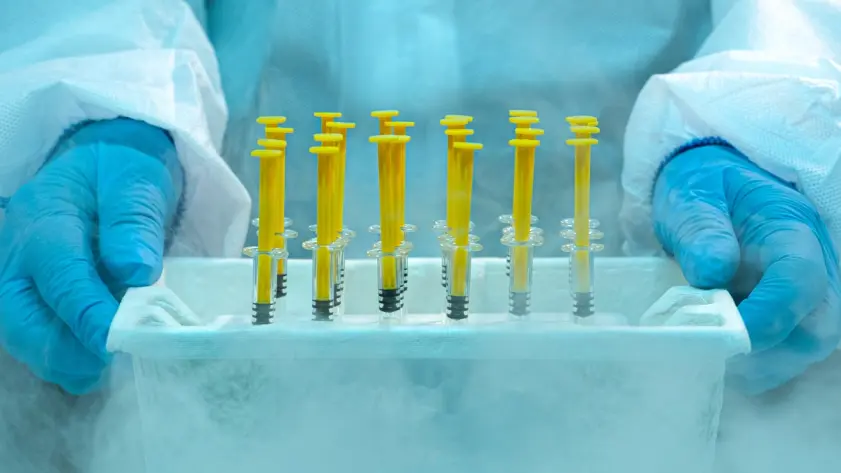 The true cost of cold storage failures - Scientist wearing protective gloves and lab suit holding frozen medical syringes in a cold storage container with visible frost and vapor.